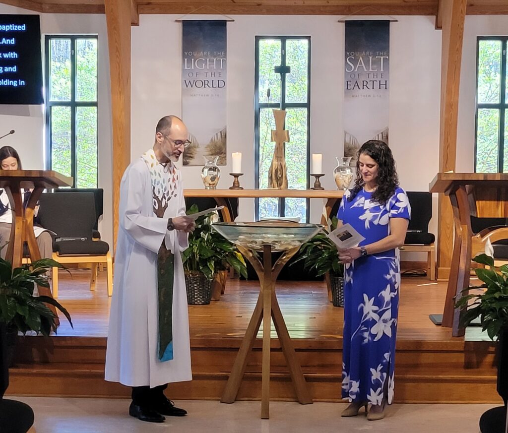 Pastor Chris presides over a baptism at Holy Covenant UCC, standing beside a woman in a blue and white floral dress at the baptismal font in the sanctuary.