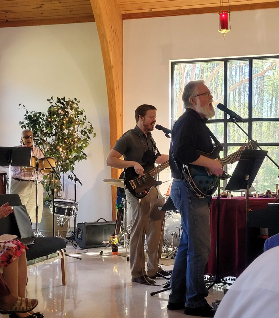 Three musicians perform during worship at Holy Covenant UCC, playing electric guitar, bass, and percussion in front of a sunlit window.