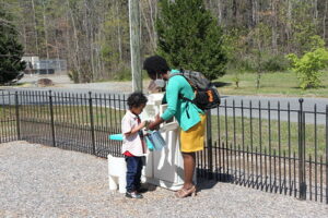 A young child holds a plastic container while a teacher kneels beside him at a playground, offering guidance and support.
