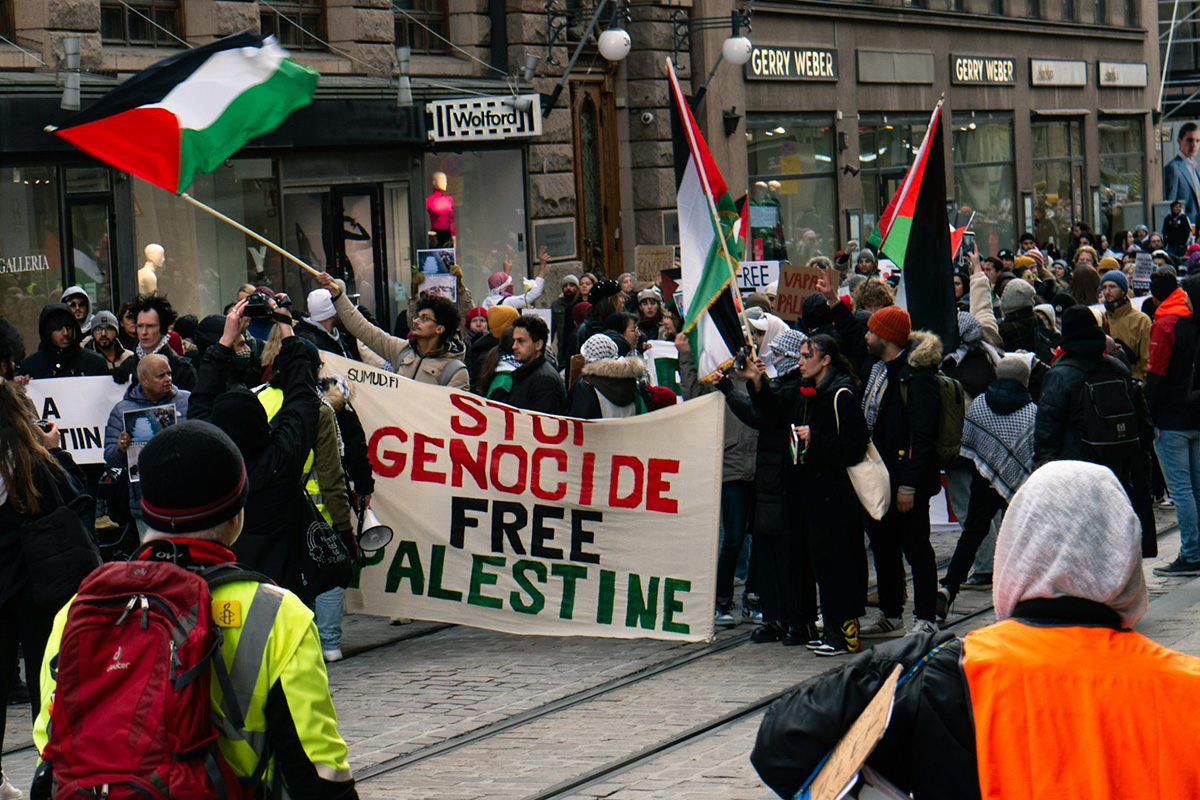 A peaceful demonstration with a banner reading 'Stop Genocide, Free Palestine' and Palestinian flags