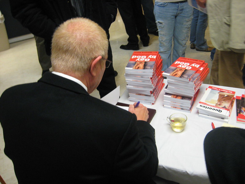 Rev. Perry signs copies of 'Gay by God' while seated at a table stacked with books.