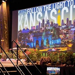 A colorful stage backdrop reads “Follow the Light to Kansas City” with a lit candle replacing the “I” in “City,” set against a vibrant skyline and blooming floral decorations in the foreground.