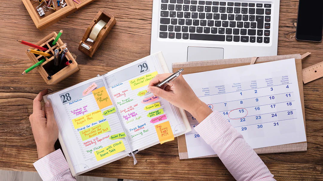 A person writing in a colorful, jam-packed paper planner with sticky notes, next to a paper calendar and laptop on a cluttered desk.