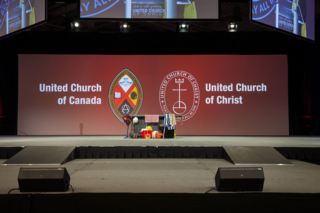 Stage with large United Church of Canada and United Church of Christ logos on red backdrop, prepared for joint worship or plenary event.